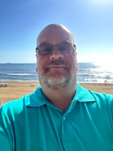 A man wearing glasses and a teal polo shirt taking a selfie near the beach with the ocean and blue sky in the background. Michael Davis
