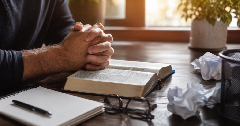 Clasped hands over an open Bible with crumpled papers nearby, representing humility, surrender, and choosing peace over winning an argument.