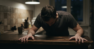 Man standing with his head down, bracing his hands against a kitchen counter, shoulders tense.